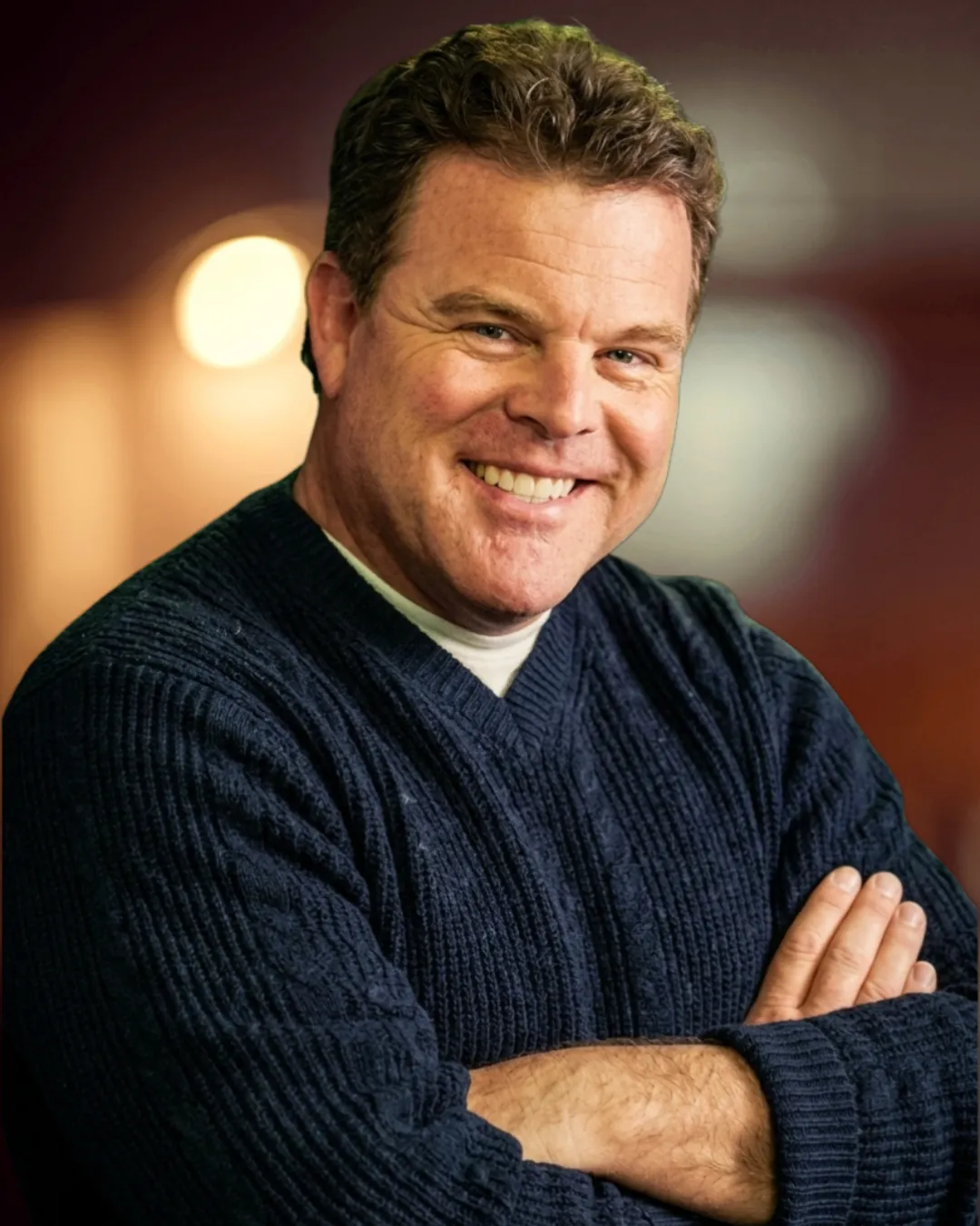 John Carney smiling with his arms crossed, wearing a sweater
